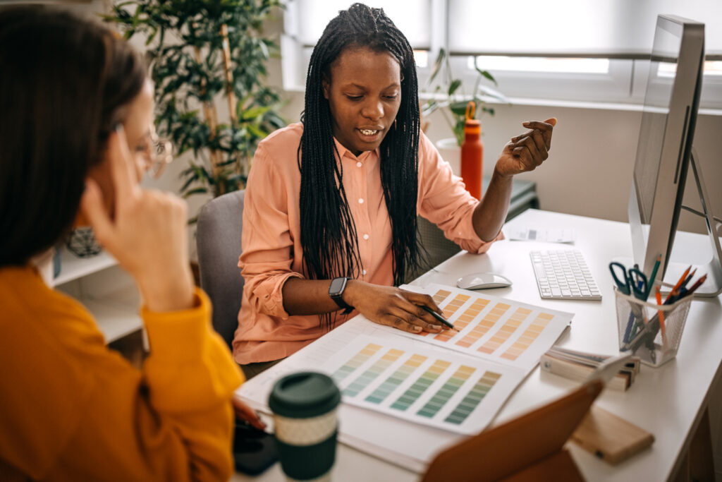 Female colleagues analyzing presentation color swatches while discussing business ideas on desk in small office