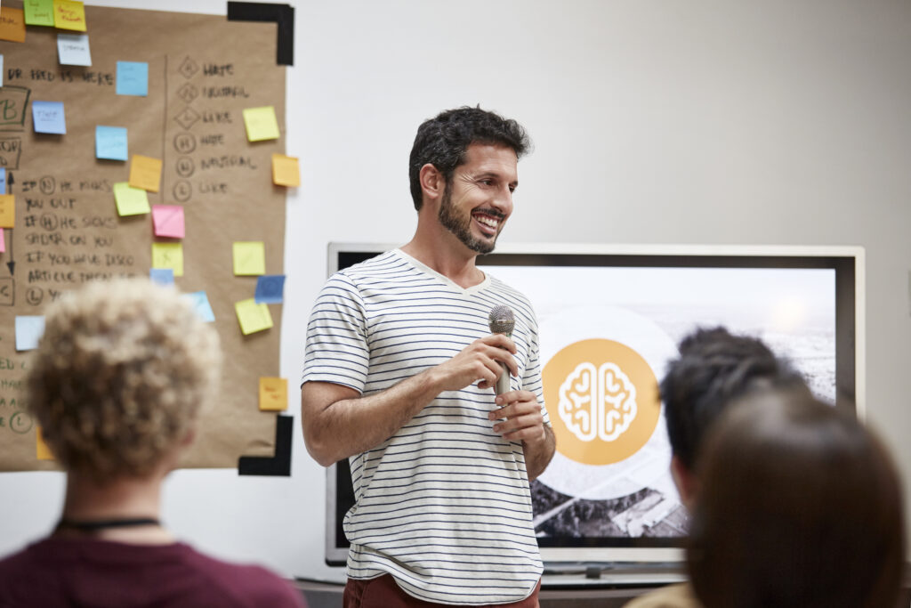 Confident businessman talking into microphone during seminar. Happy male professional is giving presentation to colleagues. He is wearing smart casuals.