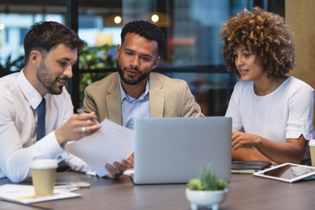 Three sales people meeting and looking at a laptop and a document. There is paperwork and other technology on the table, formal business wear.