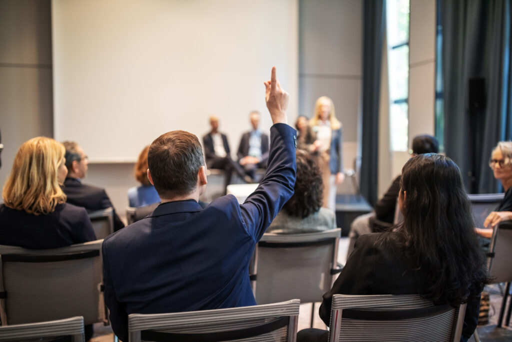 Rear view shot of a businessman raising hand to ask questions during a seminar. Professional asking query during a launch event in convention center.