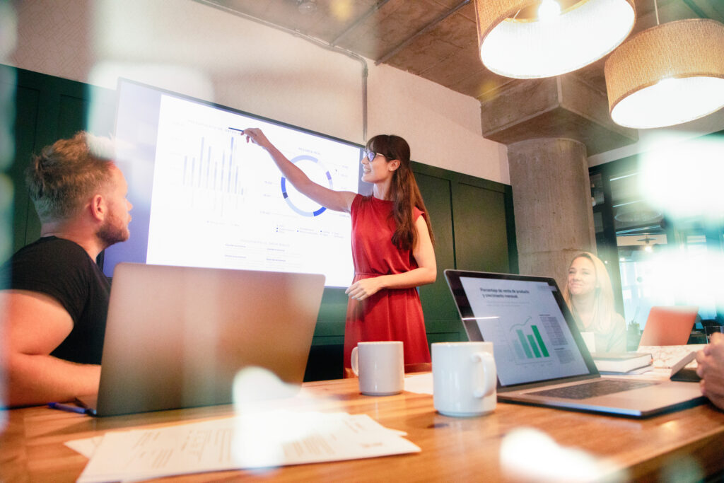 Confident business woman presents quarterly progress in meeting using a large display screen. Photographed through a window with reflections using effective presentation skills.