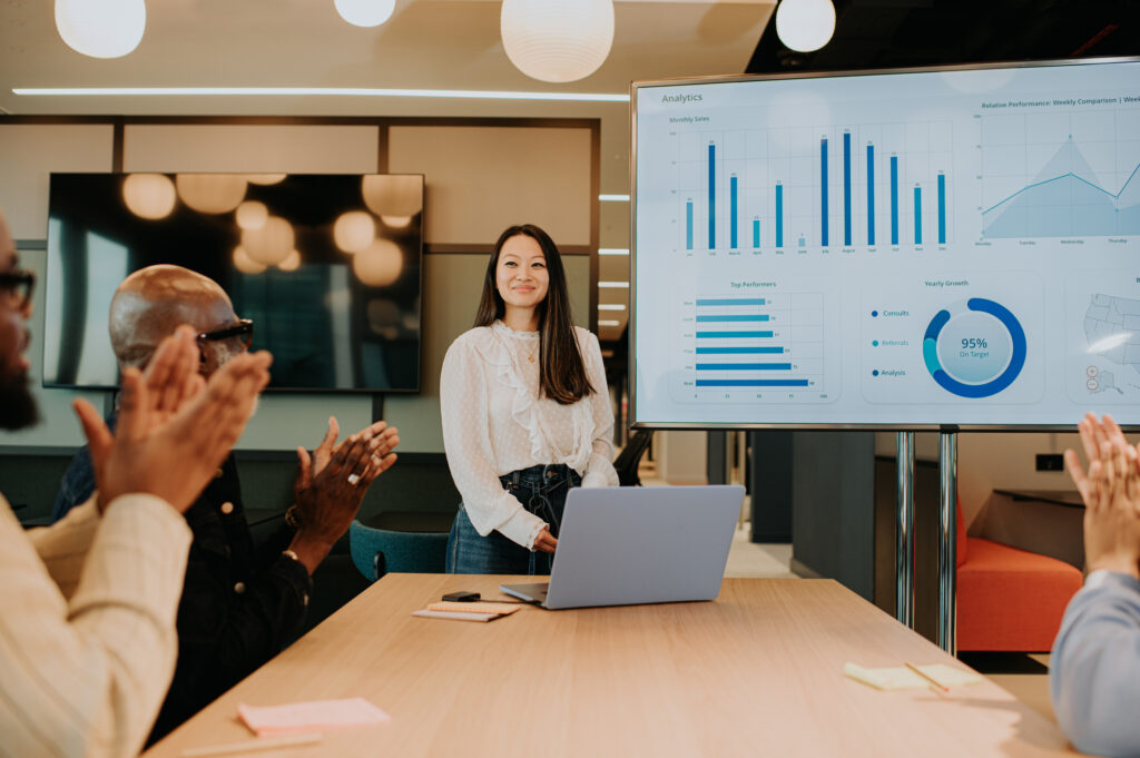 A meeting takes place in a modern office environment. A young woman has completed a successful sales deck presentation using a laptop and a huge computer monitor / television. Her colleagues giver her a round of applause for a job well done.