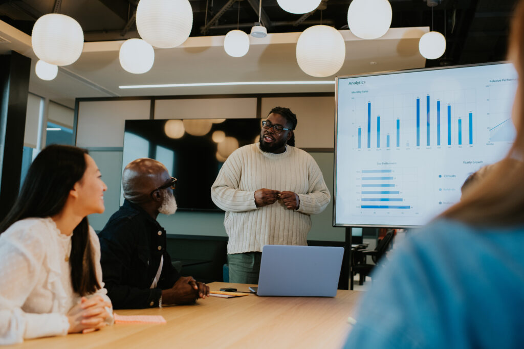 A young employee confidently stands in front of a group of colleagues in a modern office environment. He casts a screen with stats and graphs from his laptop onto a large monitor. His colleagues listen intently.