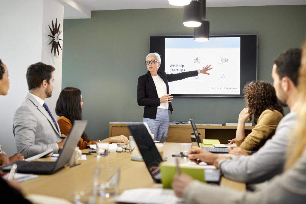 Senior businesswoman giving presentation to team. Male and female professionals are planning strategy in meeting. They are in board room at office. She is showing a business presentation theme and using the power of visual storytelling to convey the message.