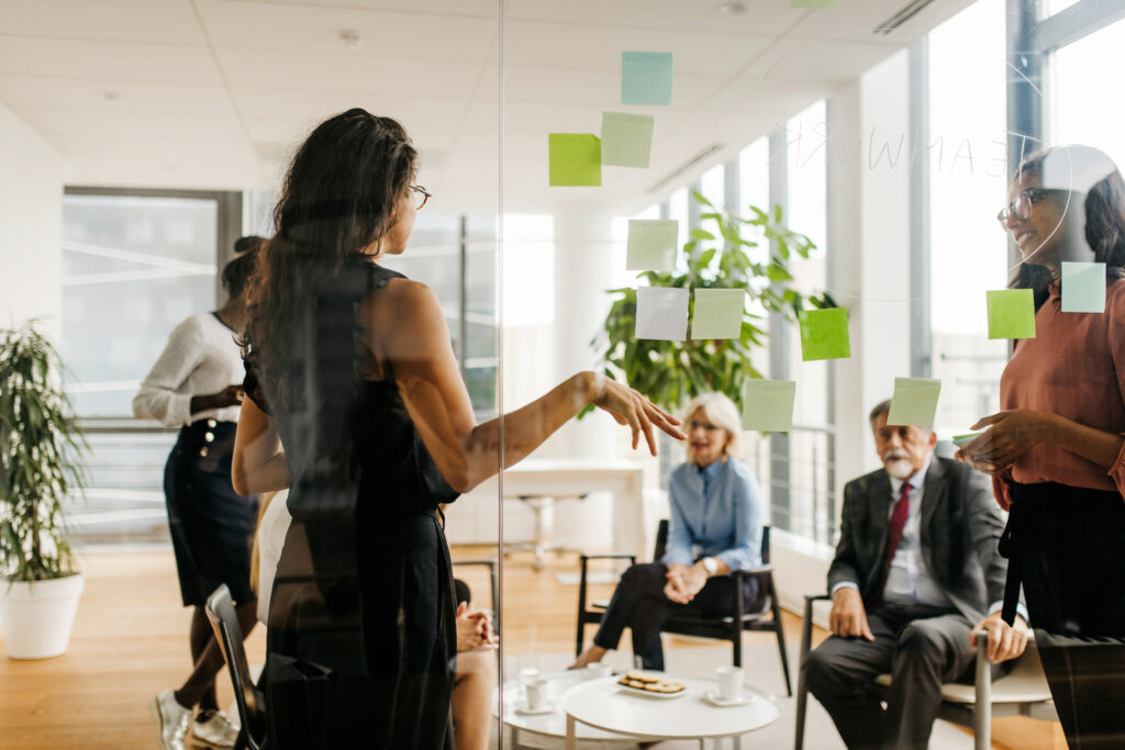 Mixed aged range of Hispanic, Indian, African, and Caucasian executives photographed through window sharing ideas and possible solutions on adhesive notes.