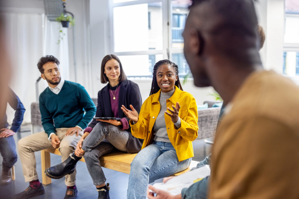 Young woman sharing her view during team building session at startup office. African woman talking with colleagues sitting in circle at a coworking office.