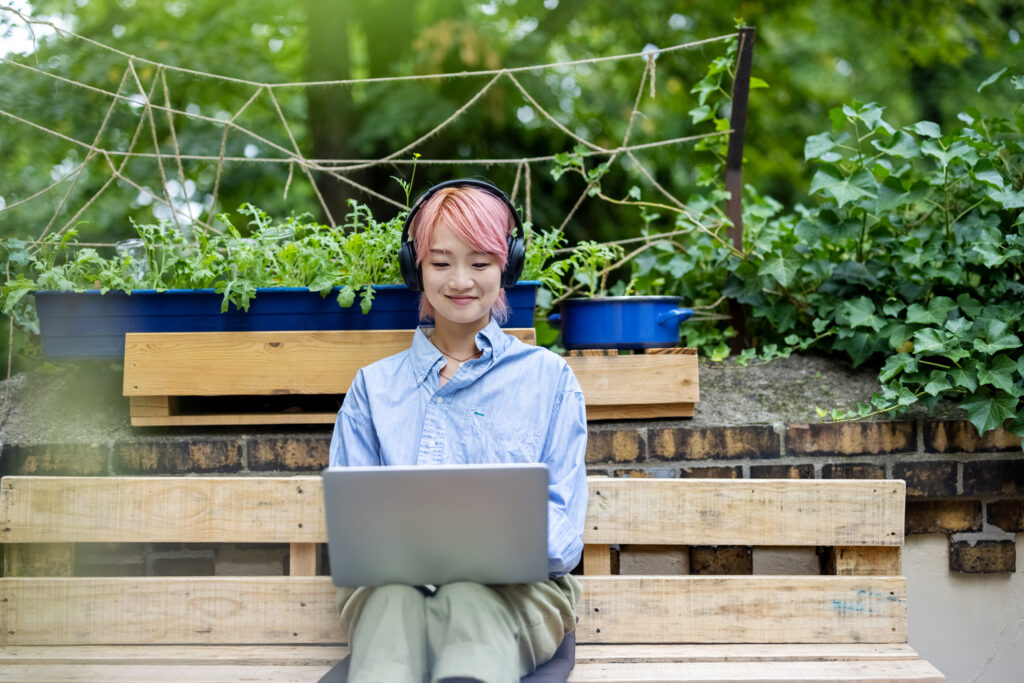 Asian woman with headphones sitting on bench outside the office and working on laptop. Female sitting on sofa using laptop, looking for presentation inspiration online.