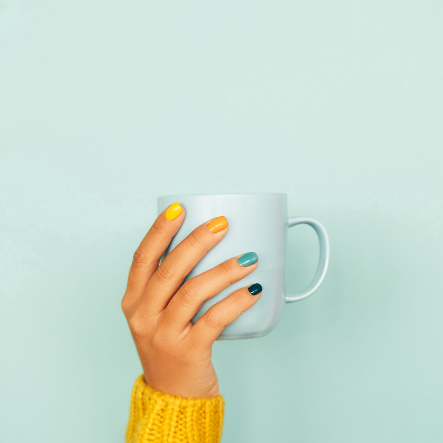 Woman hand with bright manicure and in illuminating yellow knitted sweater is holding light blue cup against light blue background. Trendy colors of the year 2021. Front view. Copy space for your design