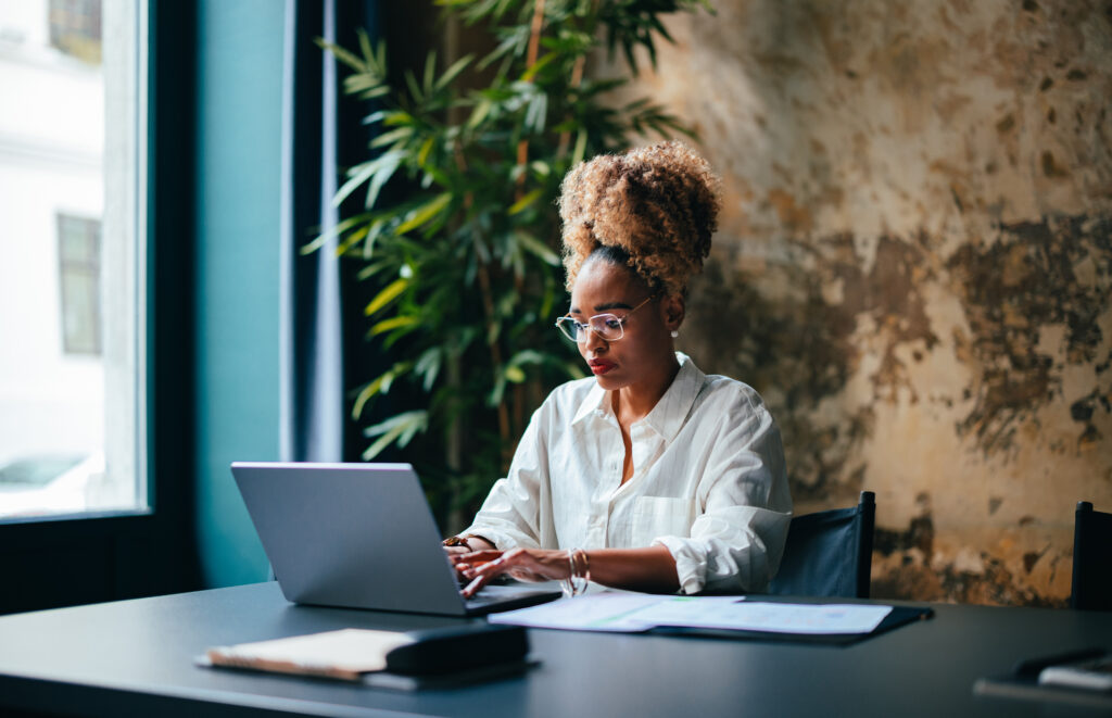 Serious African-American woman typing  business report on laptop keyboard while sitting at restaurant desk with documents.