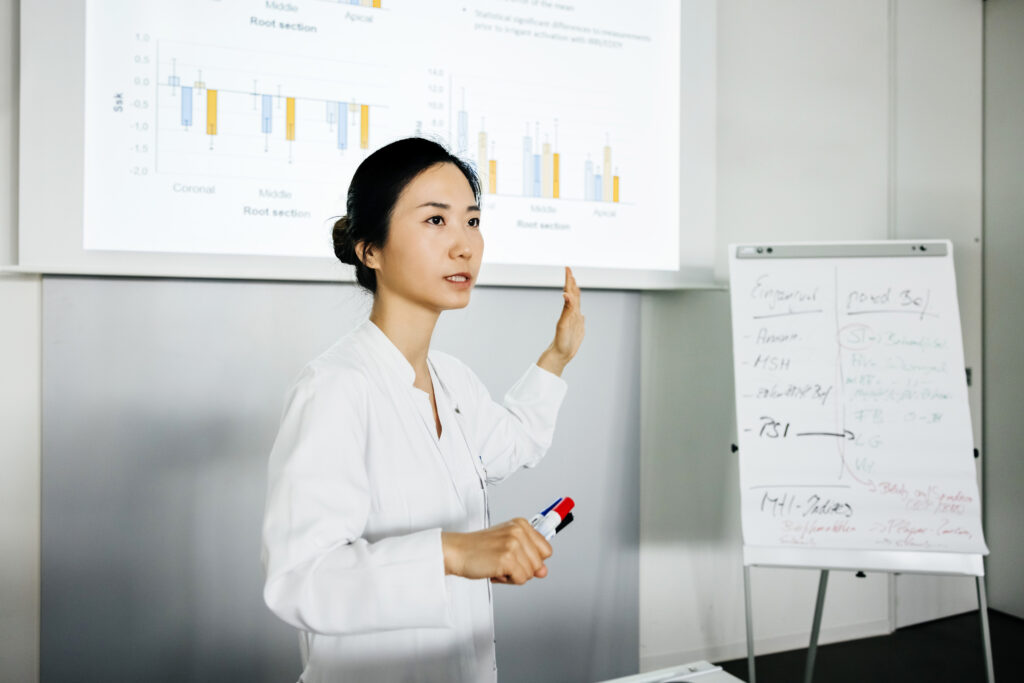 A university professor of dentistry wearing a lab coat and gesturing towards a projection on a whiteboard during a lecture.