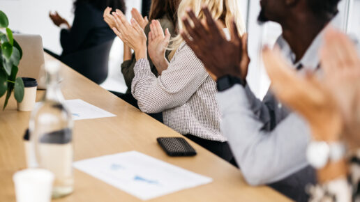 A close-up shot of co-workers clapping at a training meeting. They are sitting behind a wooden desk and are smartly dressed. Their faces are not visible. Horizontal daylight indoor photo.