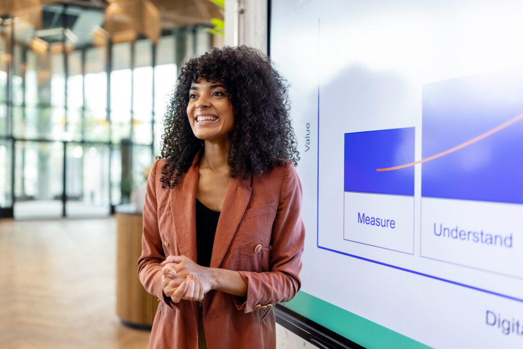 Smiling african woman giving presentation at startup. Happy female professional standing in front of a large television screen with a graph.