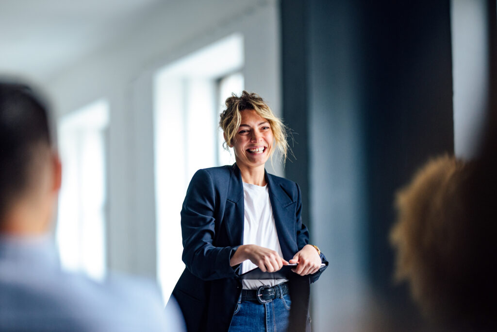 Beautiful cheerful woman in a dark blue jacket standing in front of a crowd looking down at a tablet.