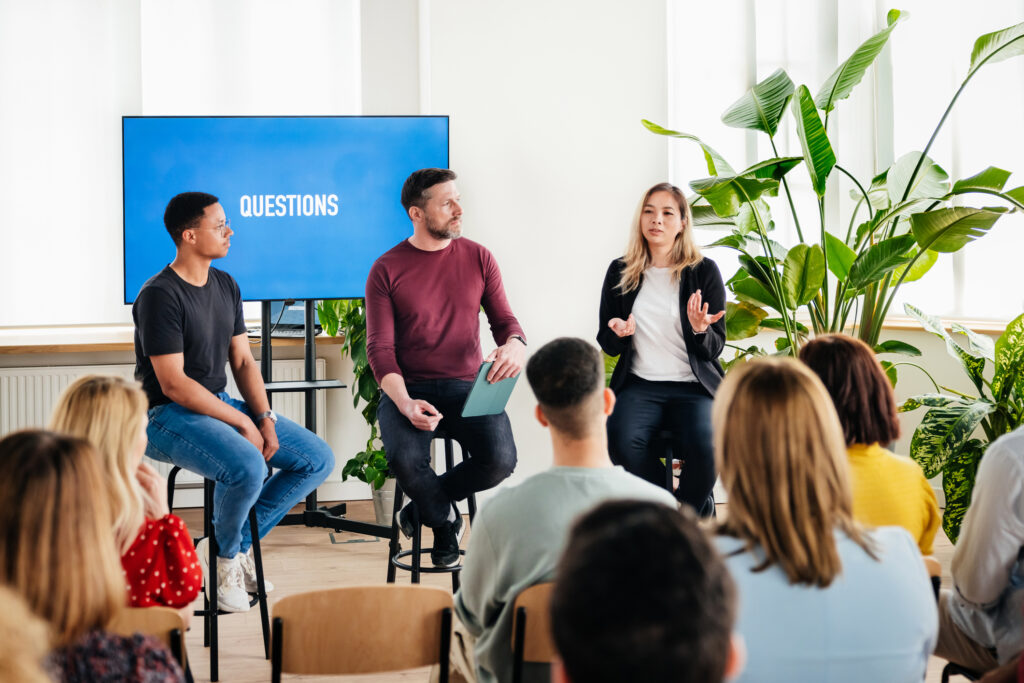 Three business employees sitting on stools and giving a seminar to a group of people.