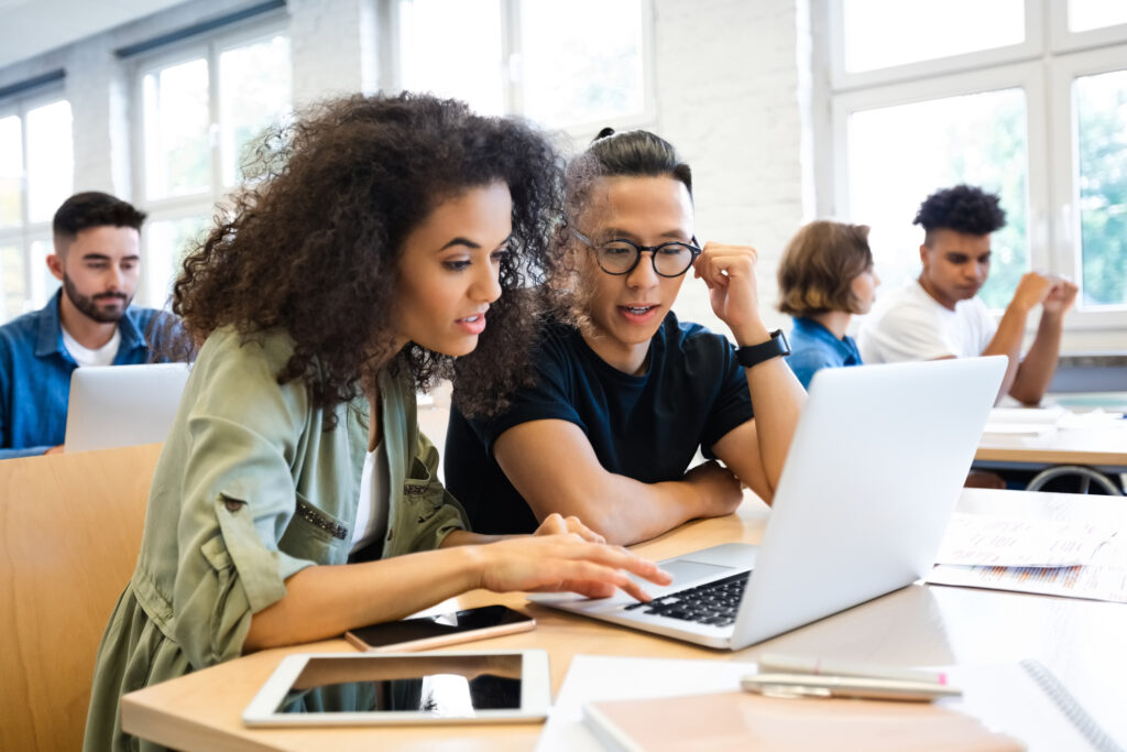 Young curly hair woman discussing with man in classroom. Classmates are learning through laptop. They are wearing casuals.