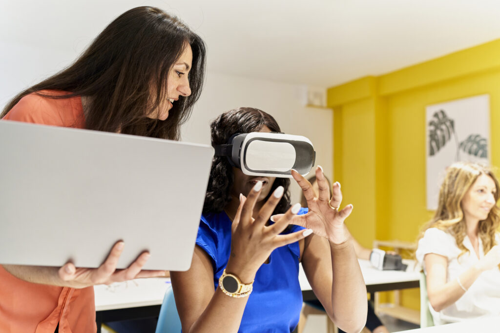 teacher in classroom holding laptop standing next to female student with hand on her man while instructing her on how to use virtual reality goggles