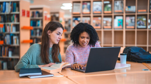 Female Hispanic professor and a Caucasian student sitting in a library studying. Sitting at a desk using laptop as a source of information.