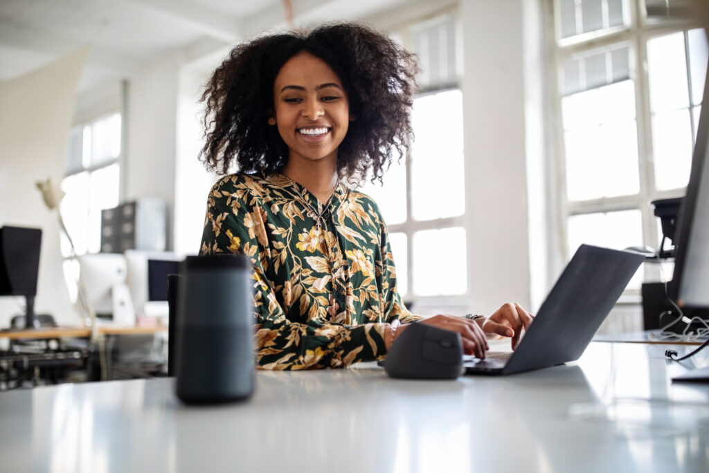 Female professional talking into a smart assistance device. Woman using virtual assistant on office table while working on laptop.