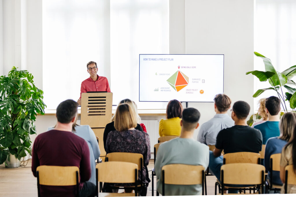 A seminar host standing behind a podium and giving a presentation to a small audience.