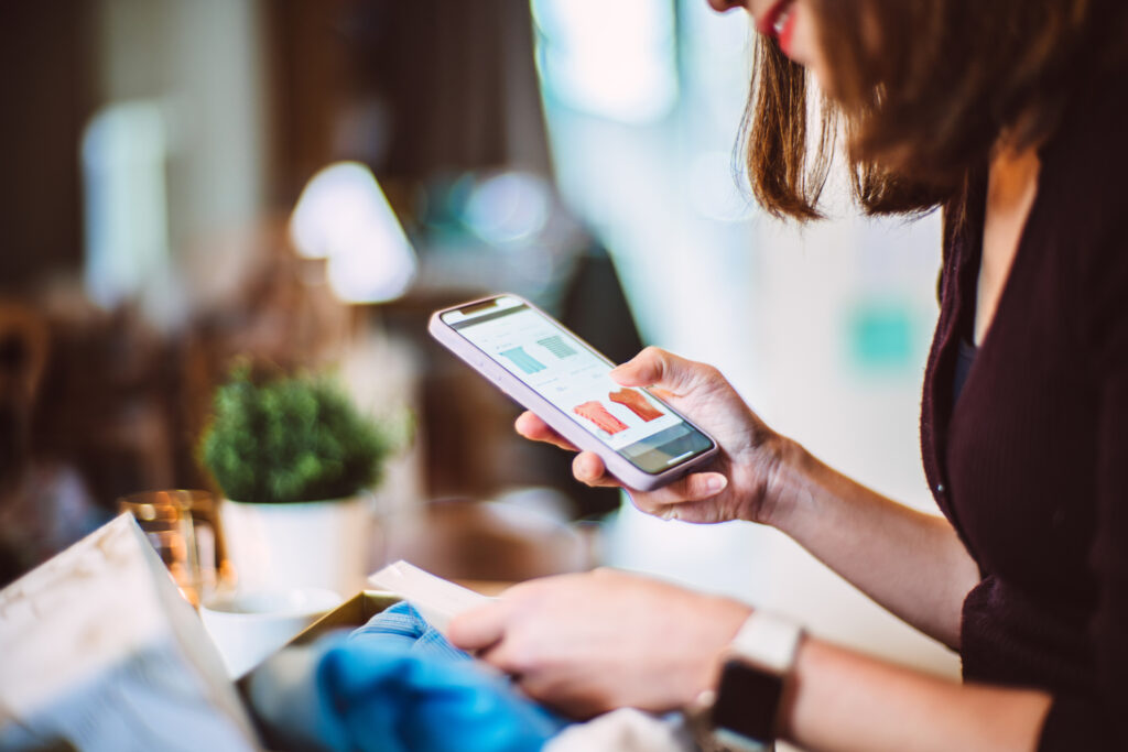 Young woman doing online shopping for clothes on smartphone at home. With delivered package from online shop putting in front of her.